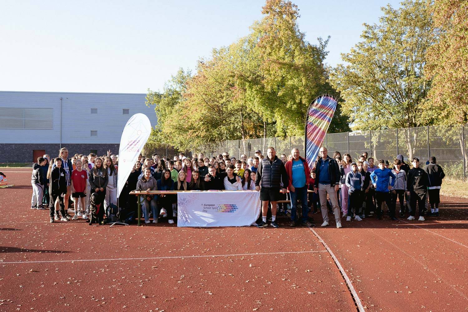 Schülerinnen und Schüler des 6. Jahrgangs stehen im Sonnenlicht auf einem Sportplatz. Ebenfalls im Bild sind eine Stempelstation, Alexander Rohr, Rainer Lose, Schulleiter Sven Schickerling, ein Banner des ESSD und Fahnen der Schule.